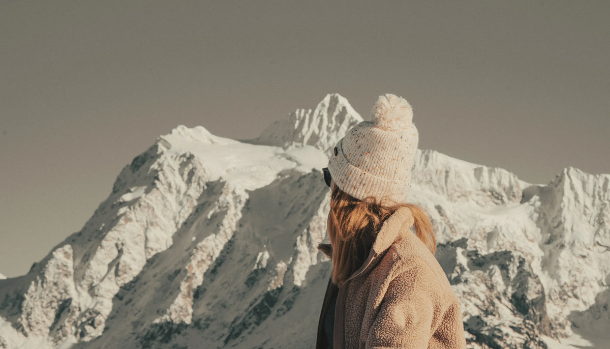 Woman in a camel teddy coat and cream knit beanie looking out over snow-covered alpine peaks