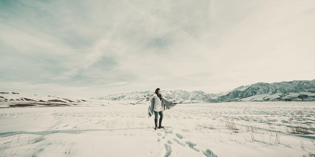 A woman in a wrap coat crossing a wide snowfield beneath distant mountains