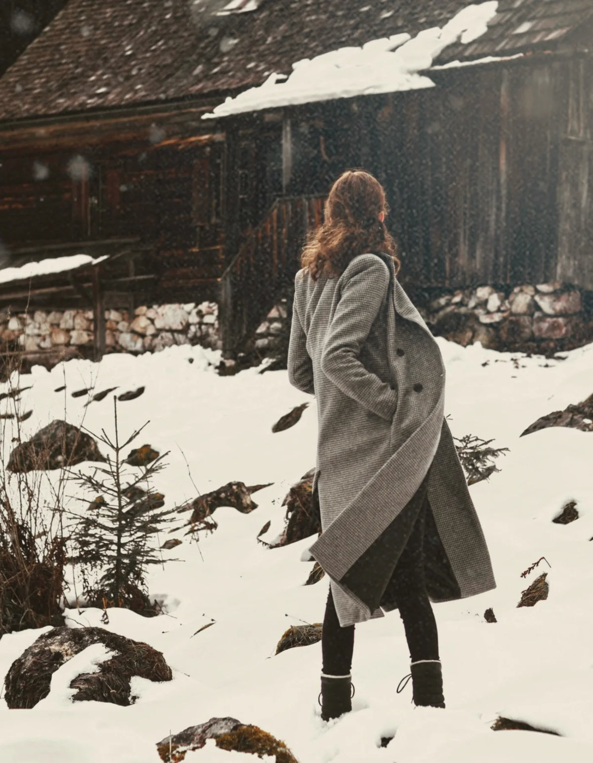 Woman in a long grey coat walking past a snow-covered wooden chalet